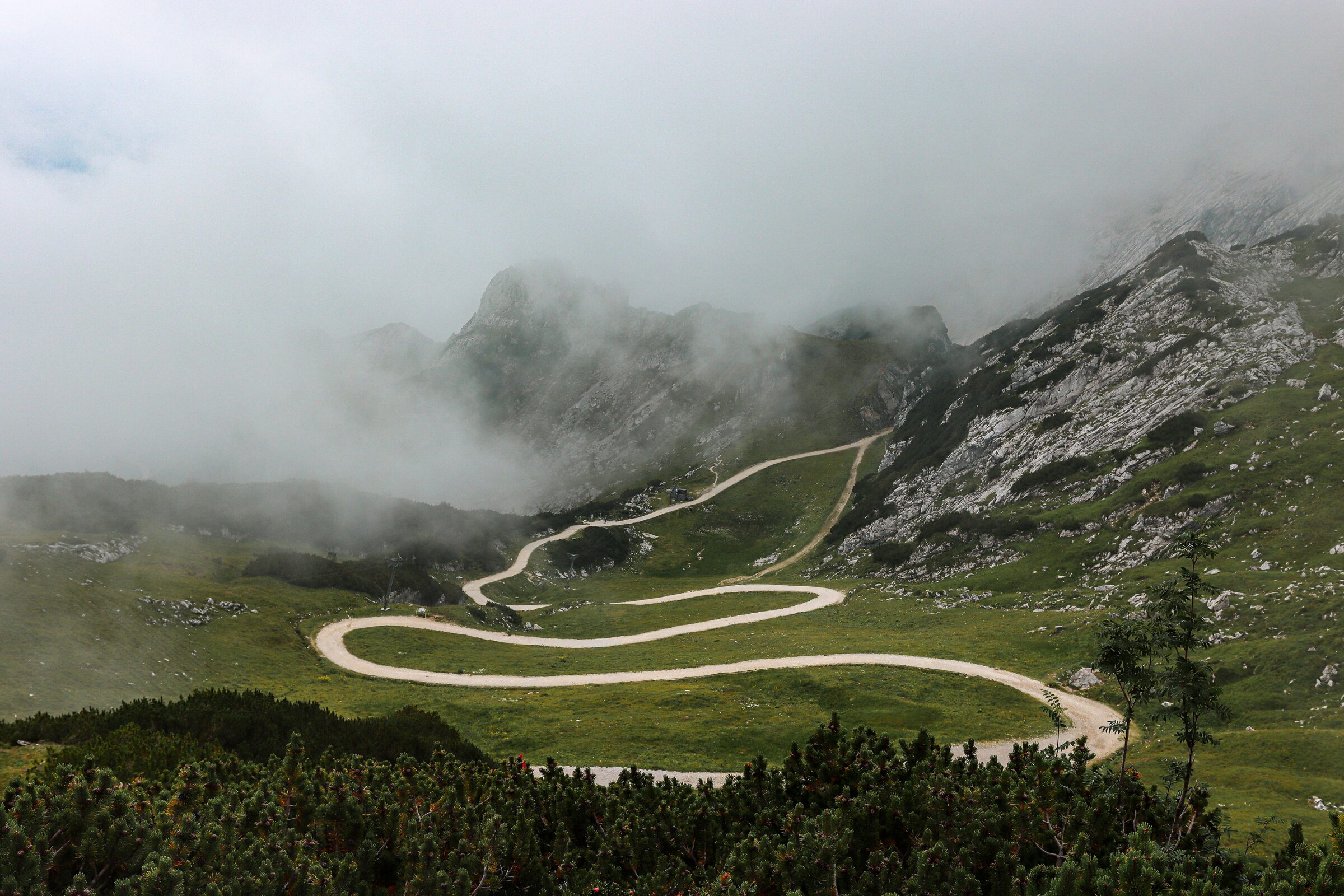 Route en épingles serpentant dans une vallée verte, brume enveloppant des sommets rocheux — un col de montagne
