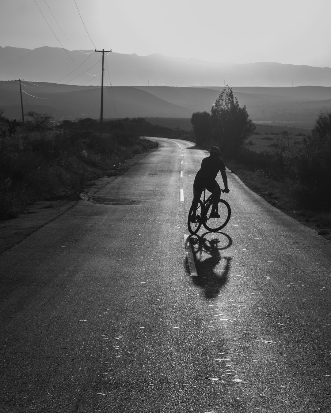 Silhouette d'un cycliste à contre-jour sur une route mouillée qui serpente vers les collines, en noir et blanc